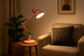 Pink desk lamp on a small round table next to a beige armchair with a dark pillow, in a cozy room setting.